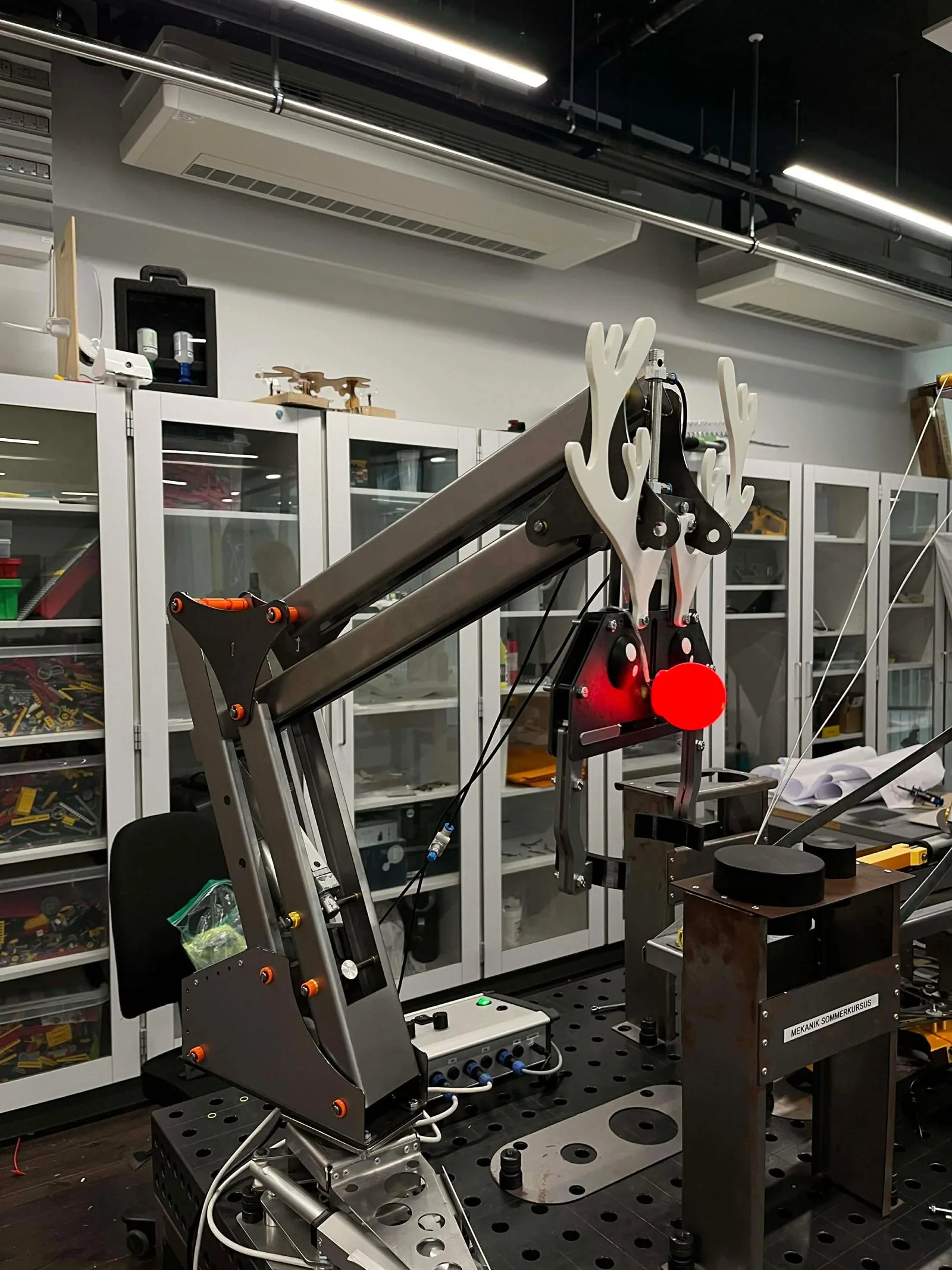 The finished robot arm in the workshop: dark steel links with orange rivet caps, a gripper with white 3D-printed antlers and a glowing red ball for a nose, above the drop-off platform.