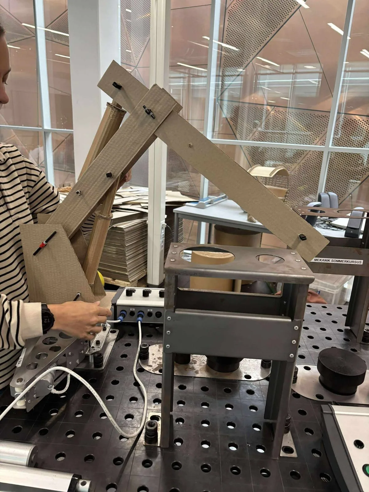 A full-size cardboard model of the arm, with pin joints and cardboard tubes standing in for actuators, mounted on the real rotating base next to the steel drop-off platform.